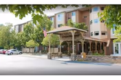 Exterior view of a multi-story senior living facility with a covered entrance, stone pillars, and an American flag. Several cars are parked in the parking lot, and trees surround the building.