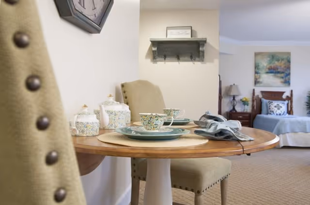 A cozy interior space featuring a small round wooden dining table set with floral teacups, saucers, and a teapot. Two beige upholstered chairs with button details are positioned around the table. In the background, there is a bed with a blue bedspread, a nightstand with a lamp and flowers, and a colorful painting on the wall. A small wall shelf with hooks is mounted above the table.