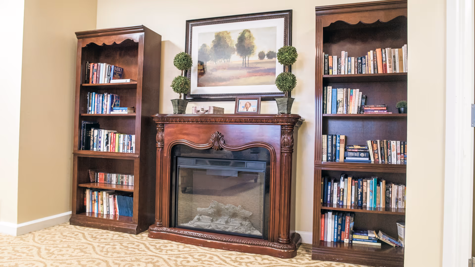 A decorative wooden fireplace mantel flanked by two bookcases filled with books, topped with a framed landscape and small potted topiaries.