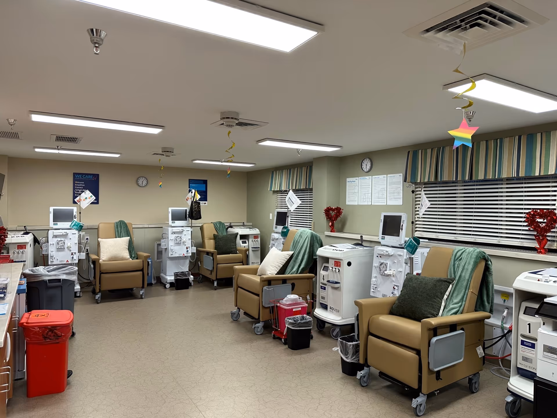 A medical treatment room with several beige recliner chairs arranged in a semi-circle, each paired with a dialysis machine. The room has overhead fluorescent lighting, windows with striped valances, and various medical supplies and waste bins. Colorful star-shaped decorations hang from the ceiling.