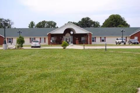 Front exterior view of a single-story senior living facility building with a green roof, brick and beige walls, a central entrance with an arched roof, two parked cars, and a large grassy lawn in the foreground.