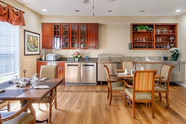 Dining room with wooden tables and chairs, hardwood floors, and a stainless-steel serving area beneath wall-mounted wooden cabinets.
