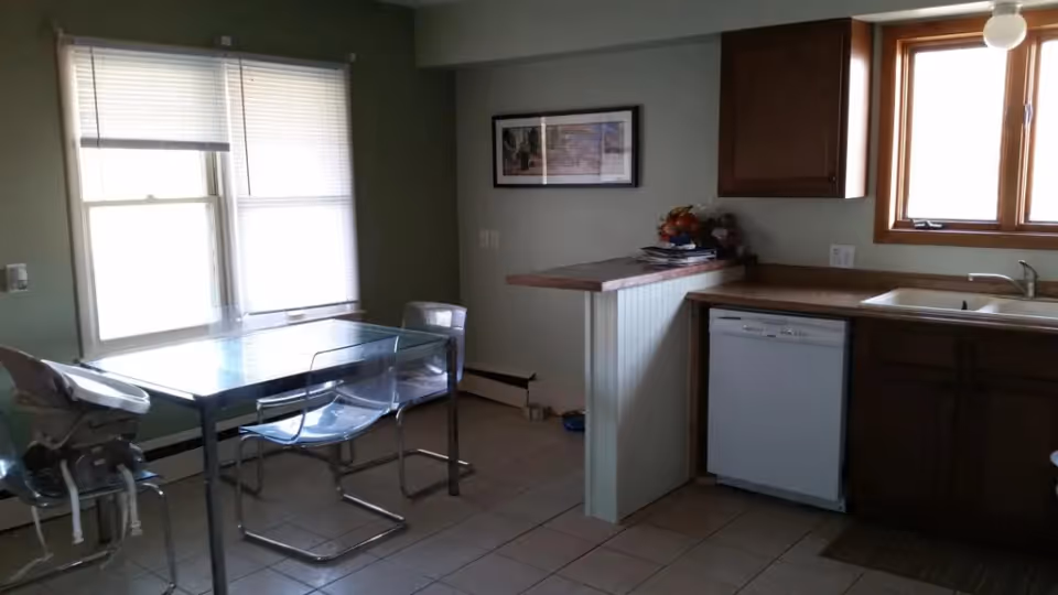 Combined kitchen and dining area with a glass dining table and clear chairs by windows, plus a countertop, sink, dishwasher, and wooden cabinets.