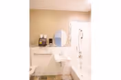 A clean bathroom with a white sink, a round mirror above it, a bathtub with a showerhead, and a grab bar on the wall. There are soap dispensers and a paper towel holder mounted on the wall.