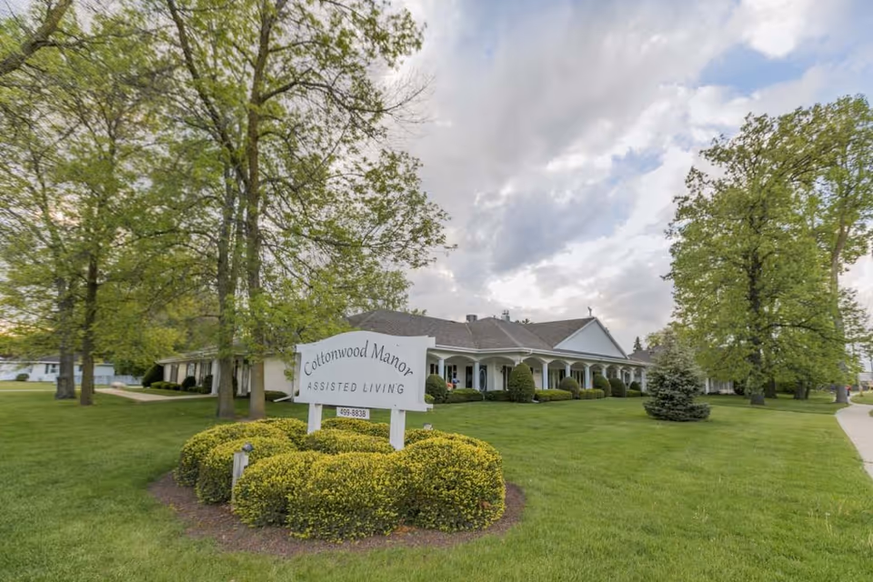 Exterior view of Cottonwood Manor Assisted Living facility with a white sign in front surrounded by neatly trimmed bushes and green grass. The building is a single-story structure with a covered porch, white columns, and several windows. Tall trees with green leaves are scattered around the property under a partly cloudy sky.