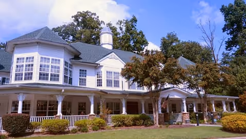 Large two-story building with white exterior and multiple windows, surrounded by trees and bushes under a blue sky with some clouds.