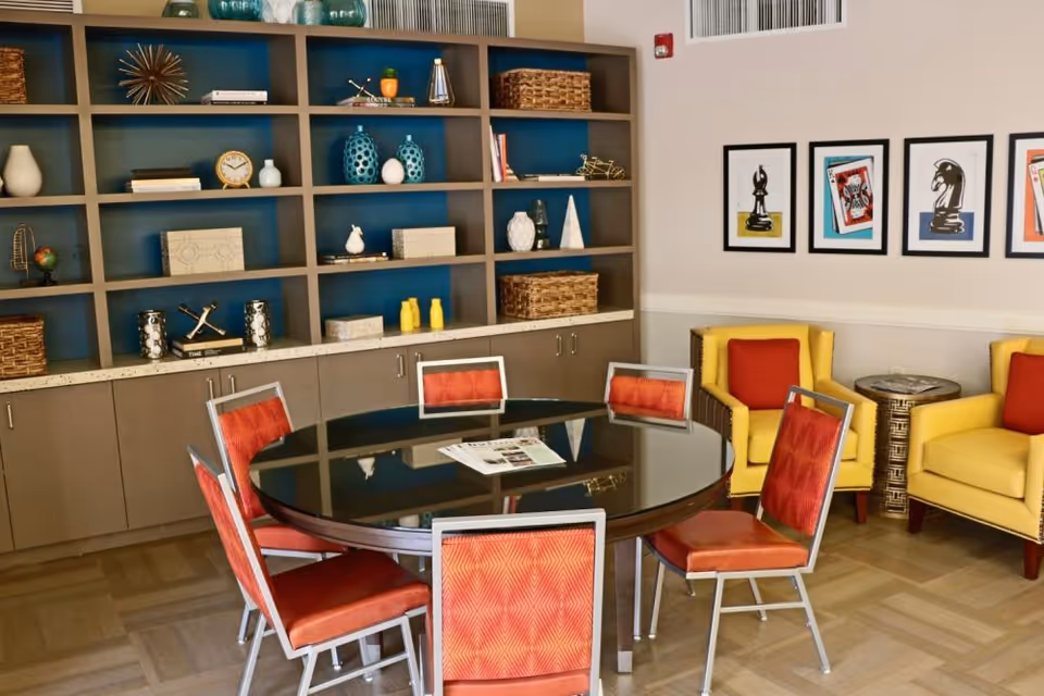 Community lounge with a round glass table surrounded by red chairs, built-in shelves with decorative items, and yellow armchairs beneath framed artwork.