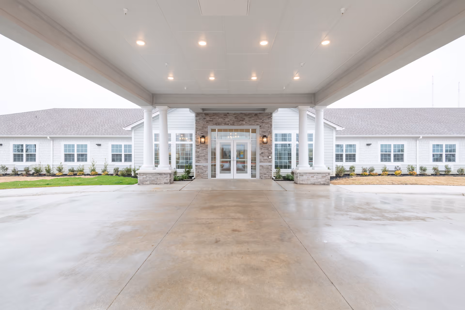Covered porte-cochere leading to the front entrance of a single-story building with white siding, columns, and large windows.