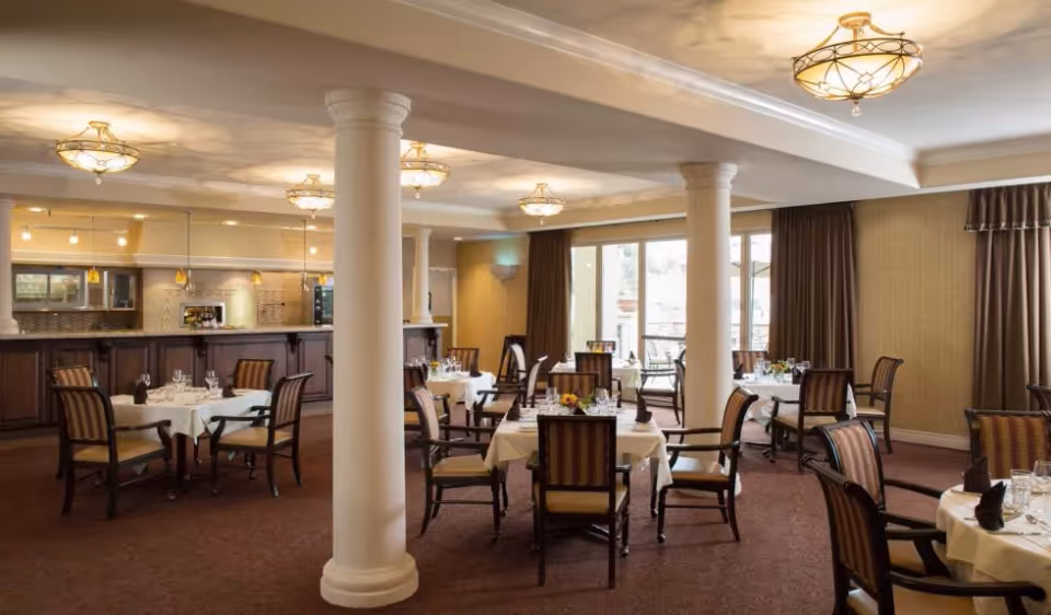 A spacious dining room with several round tables covered with white tablecloths, each set with glassware, napkins, and small floral centerpieces. The room features carpeted floors, large white columns, warm lighting fixtures on the ceiling, and large windows with brown curtains allowing natural light to enter. A counter area with pendant lights and cabinetry is visible in the background.