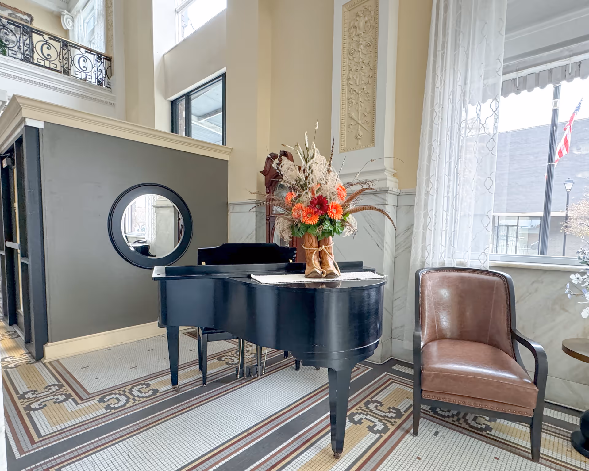 Sunlit lobby area with a black grand piano topped with a floral arrangement, a leather armchair, and decorative tiled floor near large windows.