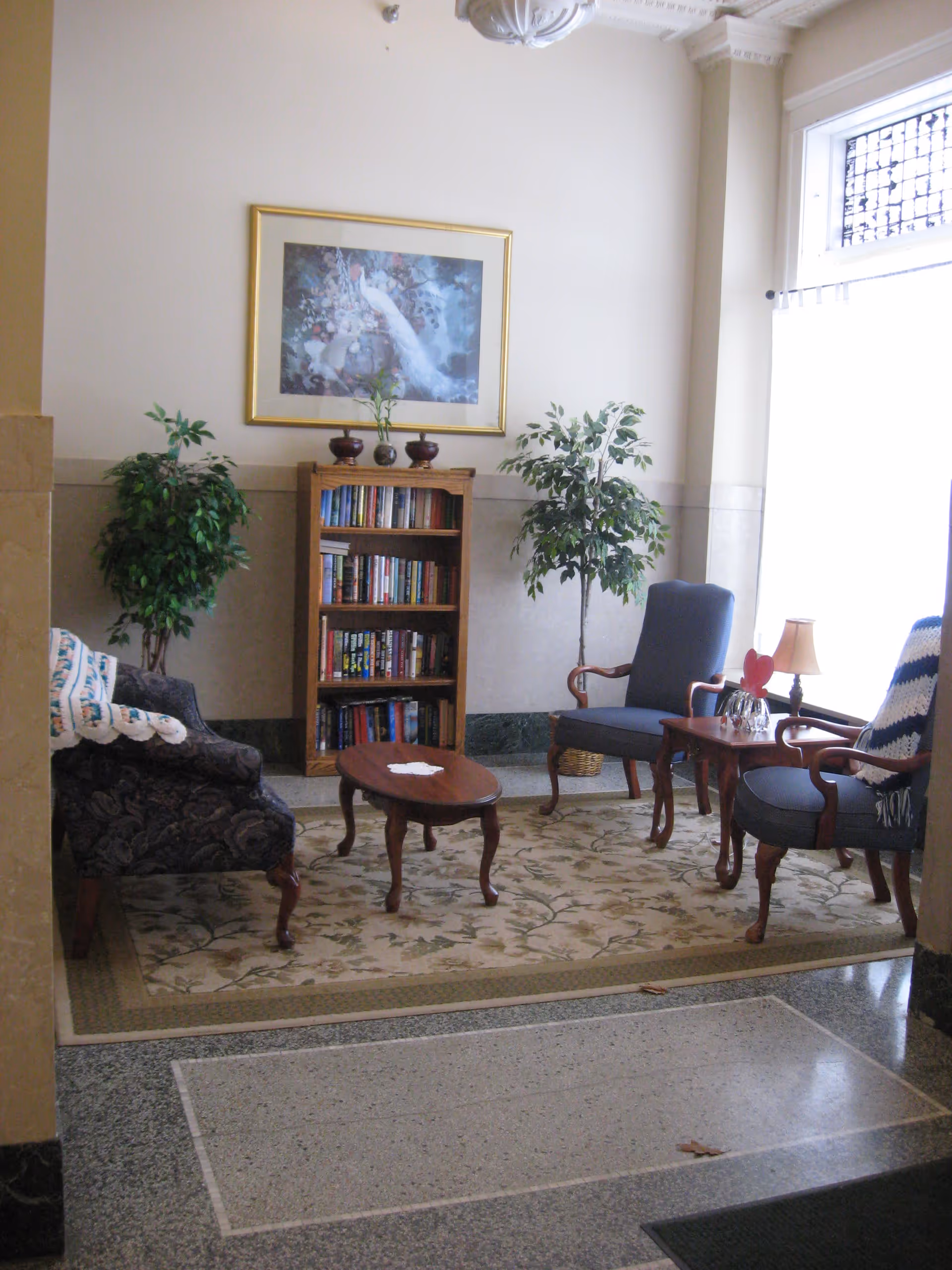 A cozy sitting area in Rumely Historic Apartments featuring two blue upholstered armchairs and one patterned armchair arranged around a small wooden coffee table on a floral area rug. Behind the chairs is a wooden bookshelf filled with books, flanked by two potted artificial plants. A framed painting of a white peacock hangs on the wall above the bookshelf. A small side table with a lamp and decorative items is next to one of the armchairs. The space is well-lit by natural light coming through a large window with a decorative grille.