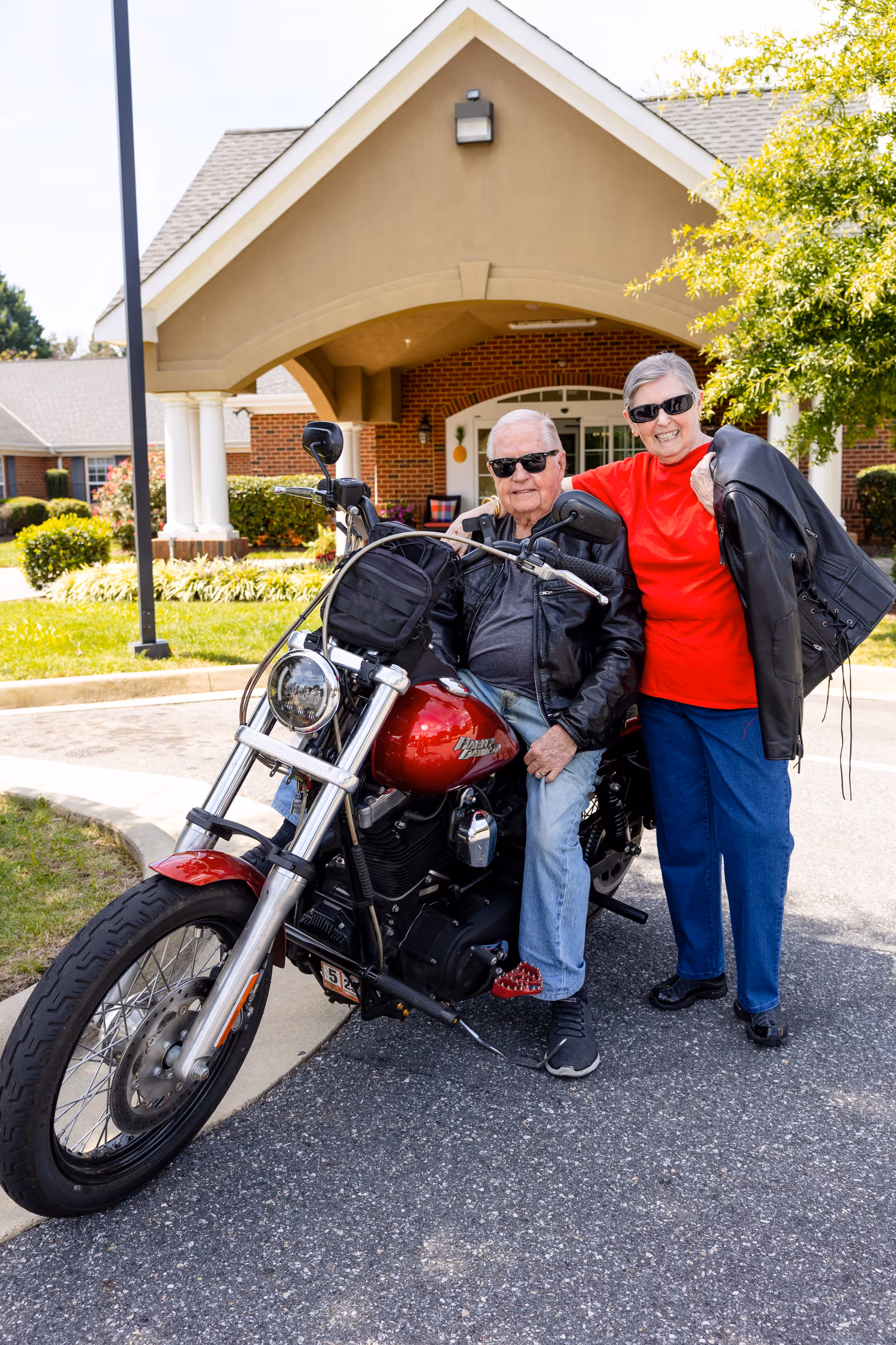 An elderly man sitting on a red motorcycle wearing sunglasses and a black leather jacket, with an elderly woman standing beside him smiling, also wearing sunglasses and holding a black leather jacket. They are outside a building with a covered entrance and greenery around.
