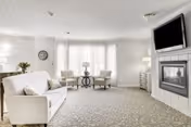 A bright and spacious living room with a white sofa, two armchairs, a round side table with a lamp, a wall-mounted flat-screen TV above a fireplace, and large windows with sheer curtains allowing natural light to fill the room.