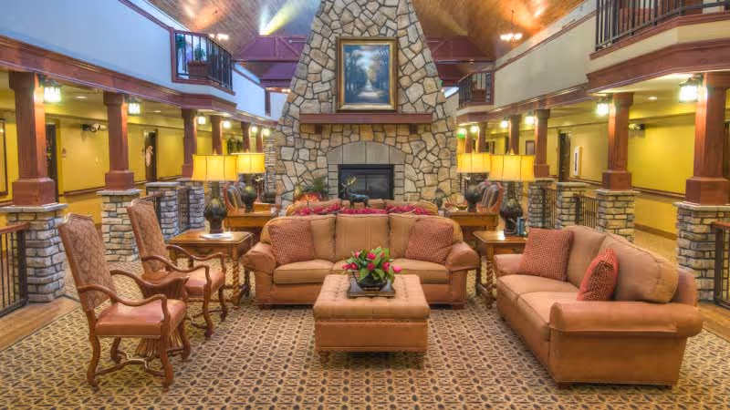 A spacious and warmly lit common area with two brown sofas, two wooden armchairs, and a cushioned ottoman arranged around a stone fireplace. The room features a high vaulted ceiling with wooden beams, a patterned carpet, and multiple table lamps providing soft lighting. A framed painting hangs above the fireplace, and the area is surrounded by a balcony with railings and stone pillars.