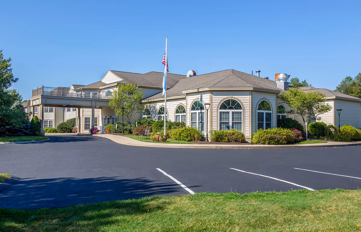 Exterior view of Benchmark Senior Living at Plymouth Crossings building with beige siding, multiple large arched windows, a covered entrance, landscaped bushes and trees, an American flag on a flagpole, and a clear blue sky.