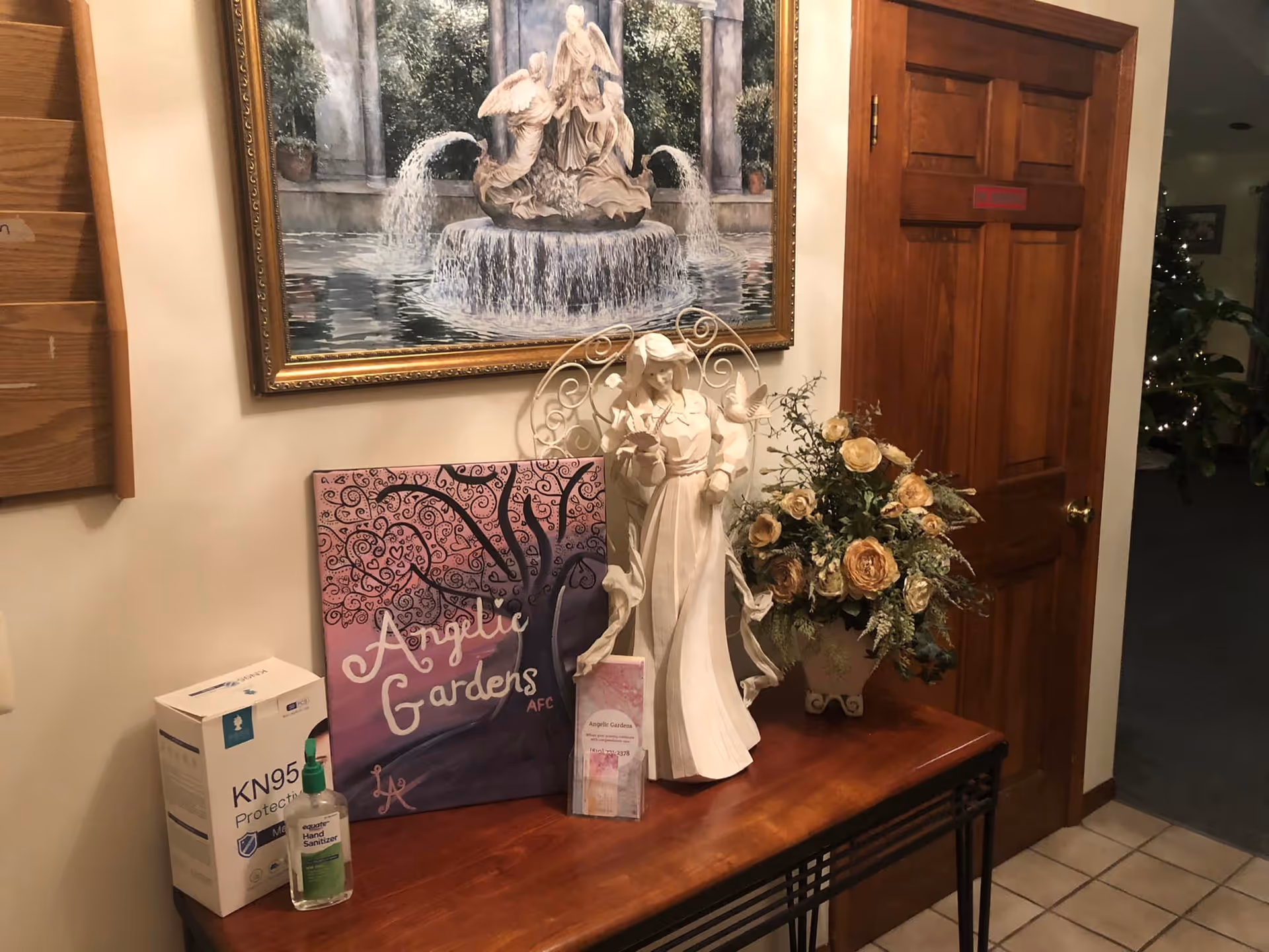 A wooden table against a wall with a decorative white statue of a woman holding birds, a bouquet of artificial flowers in a vase, a box of KN95 protective masks, a bottle of hand sanitizer, and a purple sign reading 'Angelic Gardens'. Above the table is a framed painting of a fountain with angel statues. To the right is a wooden door with a red sign, and a hallway with plants and a Christmas tree with lights is visible in the background.