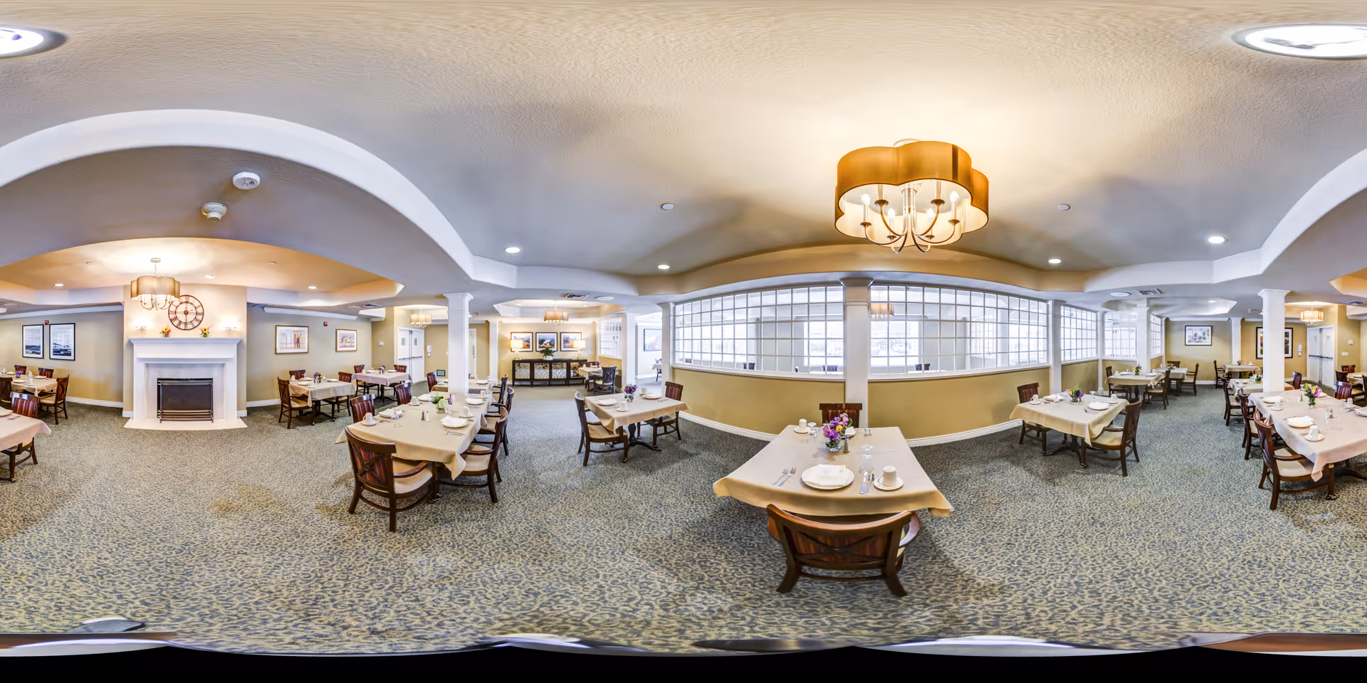 A spacious dining room in a senior living facility with multiple tables covered with beige tablecloths, set with plates, cups, and silverware. The room features a carpeted floor, a white fireplace with a large clock above it, framed artwork on the walls, and large windows with glass blocks allowing natural light. Warm ceiling lights and chandeliers provide additional lighting.