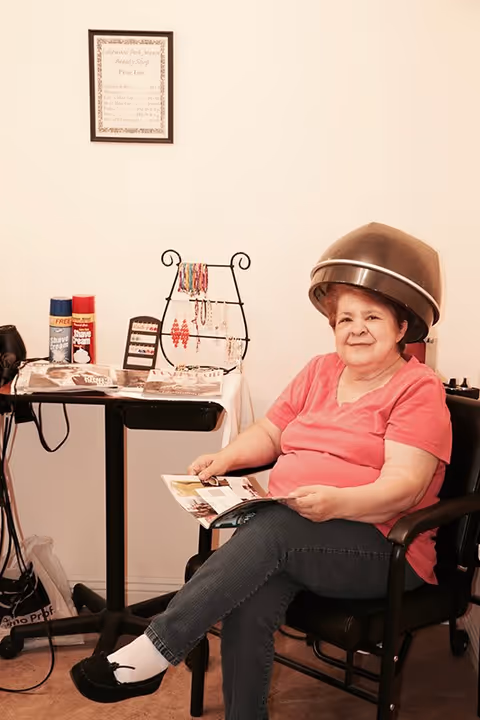 An elderly woman sitting under a hair dryer in a beauty shop area, wearing a pink shirt and blue jeans, holding a magazine. A small table next to her displays hair products and jewelry.