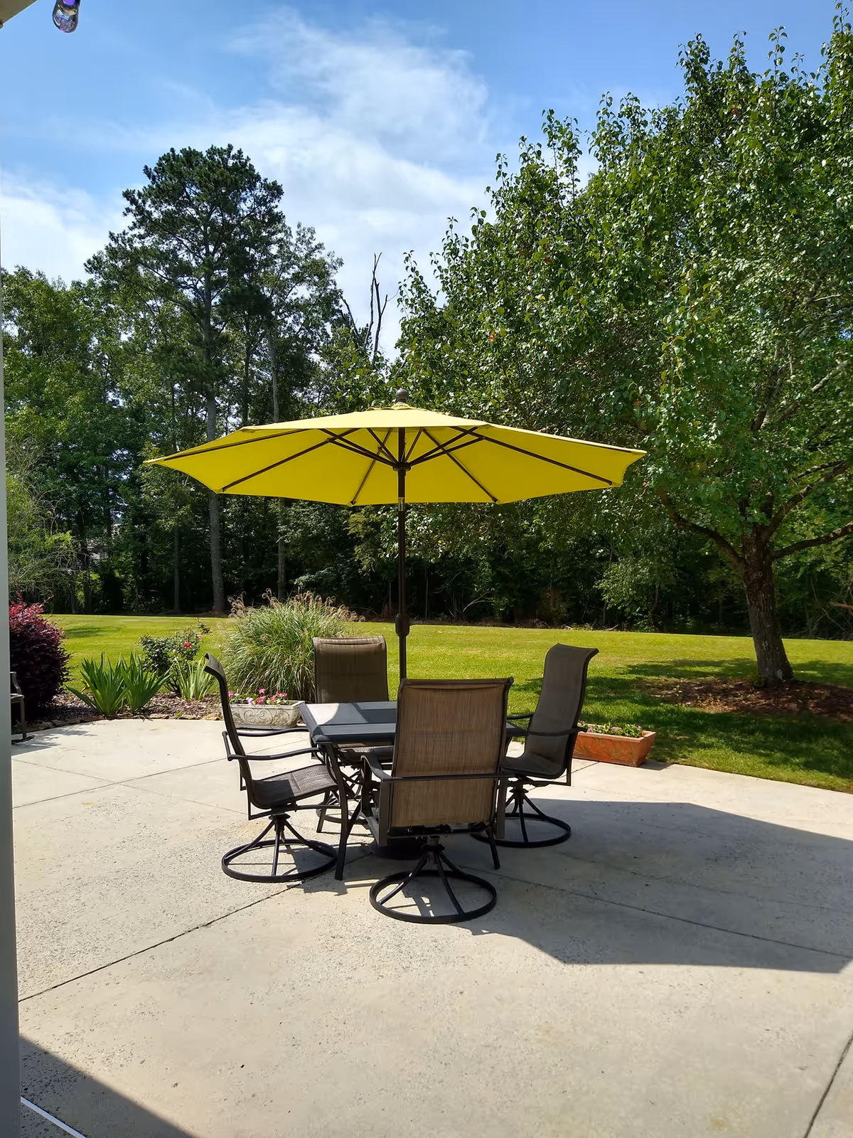 Outdoor patio area with a square glass table surrounded by four brown mesh chairs. A large yellow umbrella is open above the table providing shade. The patio is concrete and surrounded by green grass, trees, and shrubs under a partly cloudy blue sky.