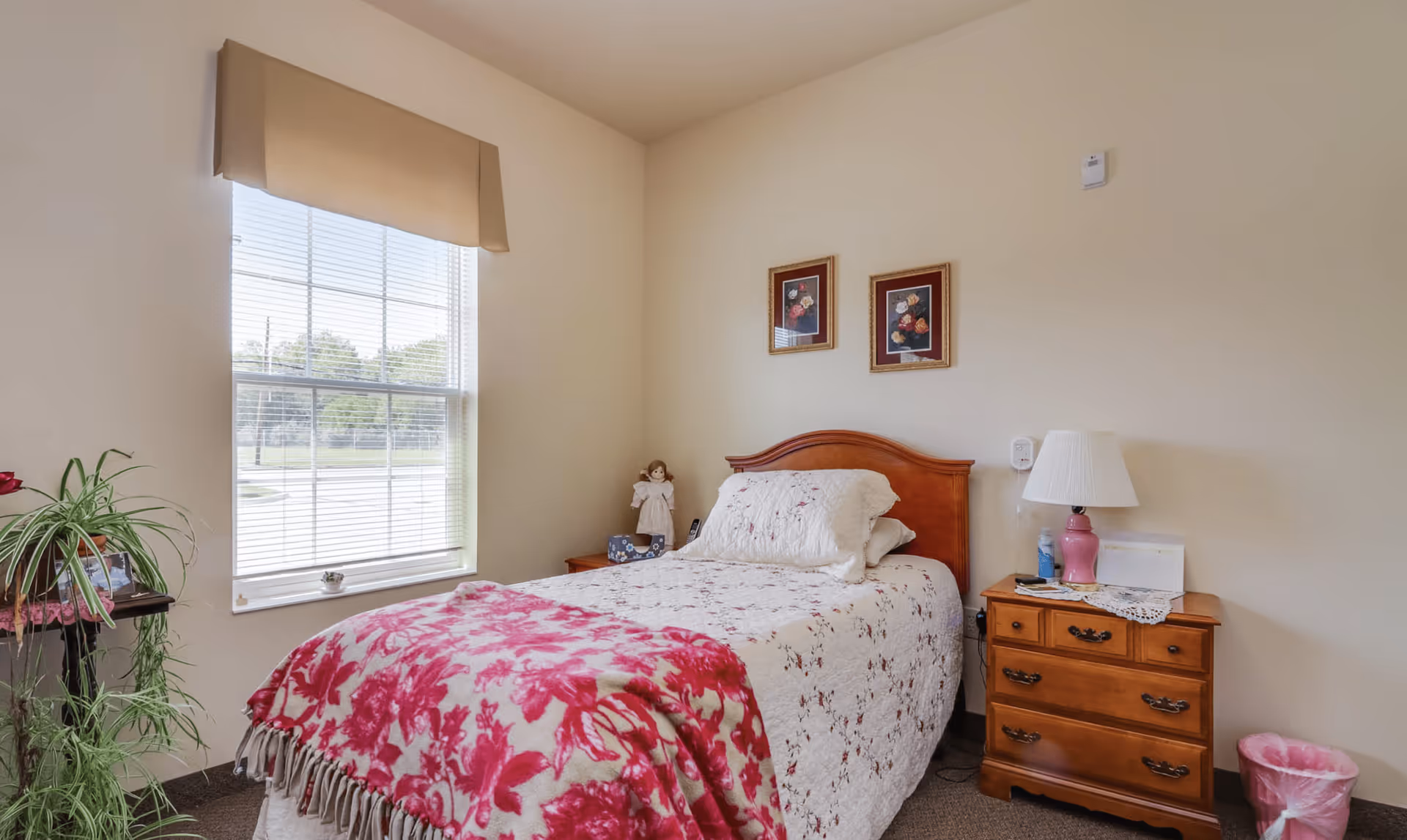A cozy bedroom with a single bed covered in a white quilt with floral patterns and a pink floral blanket. There is a wooden headboard and a wooden nightstand with a pink lamp, a doily, and some items on it. Two framed floral pictures hang on the beige wall above the bed. A window with blinds and a beige valance lets in natural light. A green plant is on a small table near the window, and a pink wastebasket is on the floor next to the nightstand.