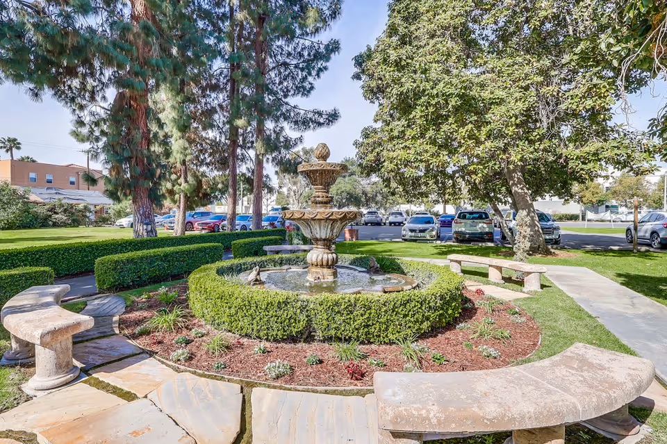 A landscaped outdoor garden area featuring a circular stone water fountain surrounded by neatly trimmed hedges and flower beds. Stone benches are arranged around the fountain on a paved area. Tall trees and parked cars are visible in the background under a clear sky.