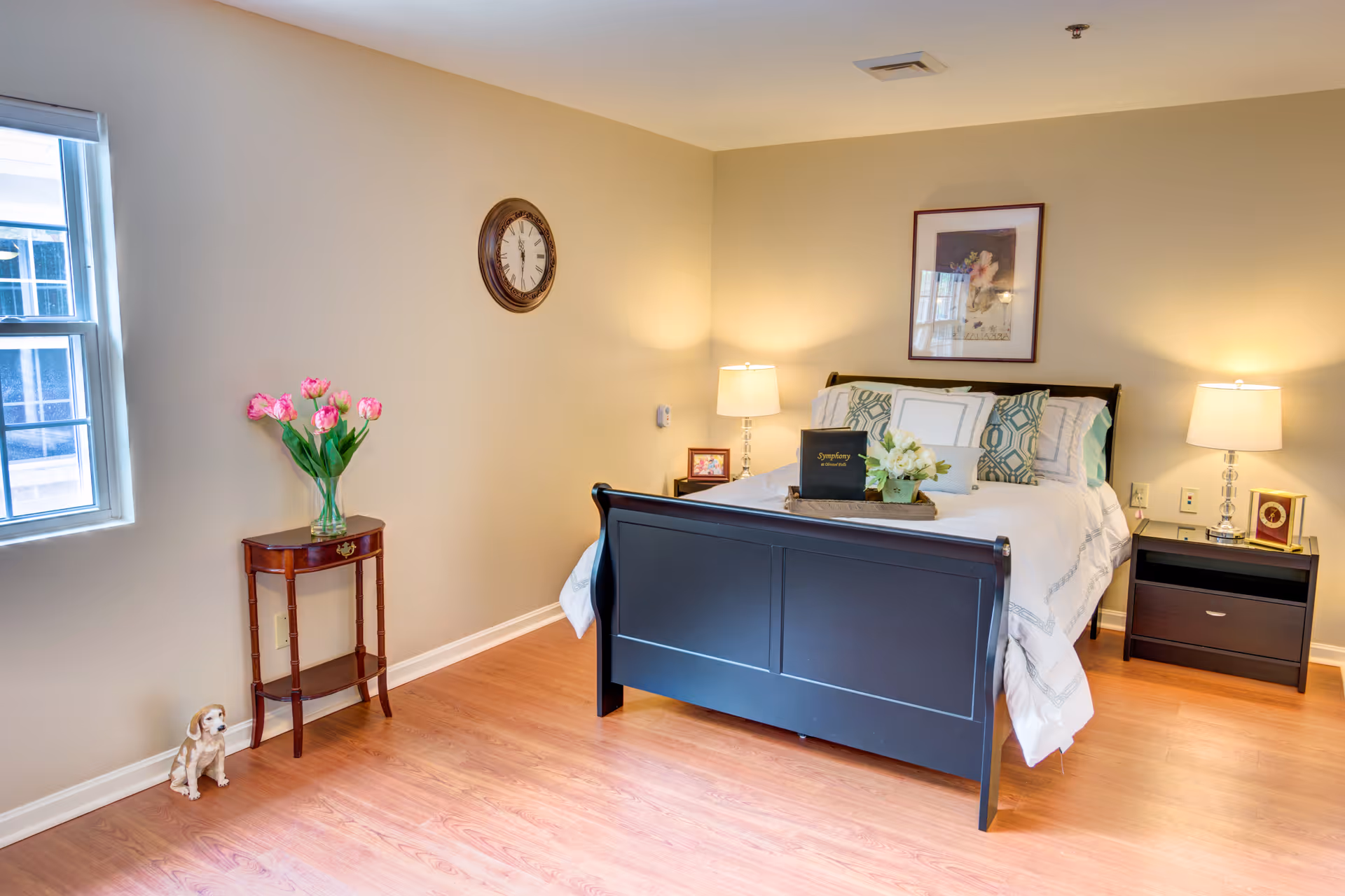 A cozy bedroom with a dark wooden bed frame, white bedding, and decorative pillows. Two bedside tables with lamps flank the bed. A framed picture hangs above the bed. To the left, there is a small wooden table with a vase of pink flowers and a window letting in natural light. A small dog figurine sits on the floor near the table. The room has light-colored walls and wooden flooring.