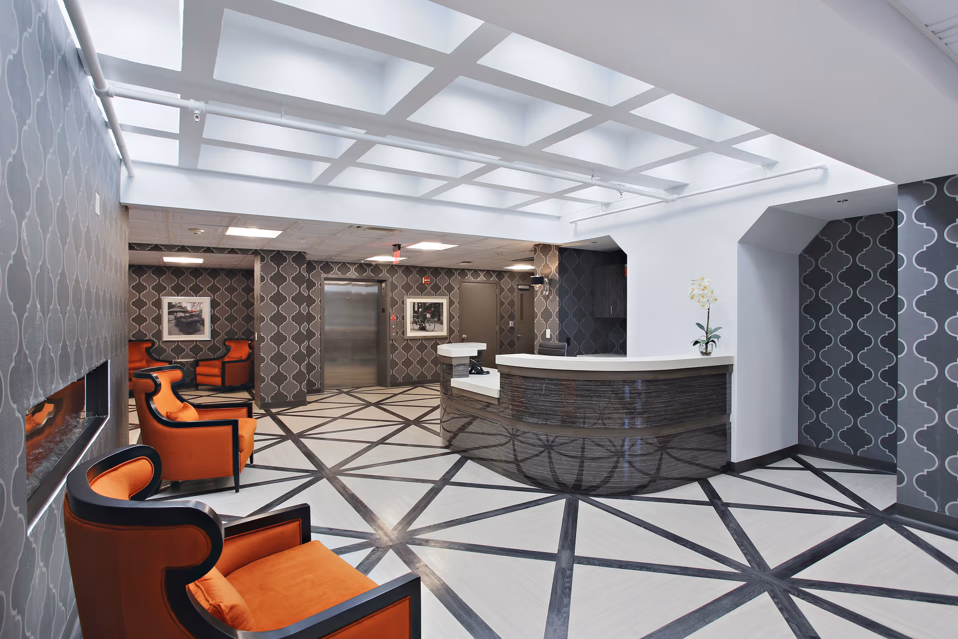 Modern reception lobby with a curved front desk, geometric patterned floor, and orange seating under a bright coffered ceiling.