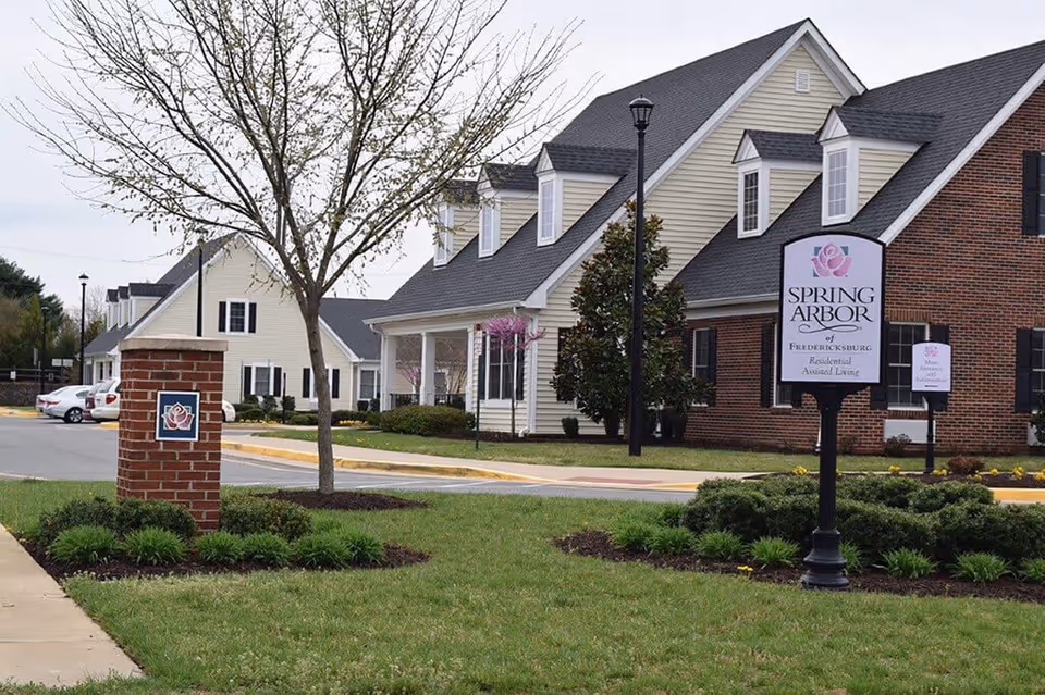Exterior view of Spring Arbor of Fredericksburg, a residential assisted living facility, showing a landscaped lawn with a tree, a brick pillar with a rose emblem, and a sign with the facility's name in front of beige and brick buildings with black shutters.