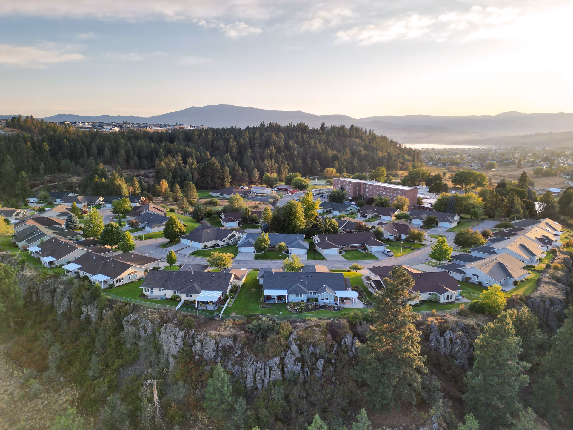Aerial view of a residential community on a rocky bluff with single-story homes, a larger facility building, and forested hills in the background.