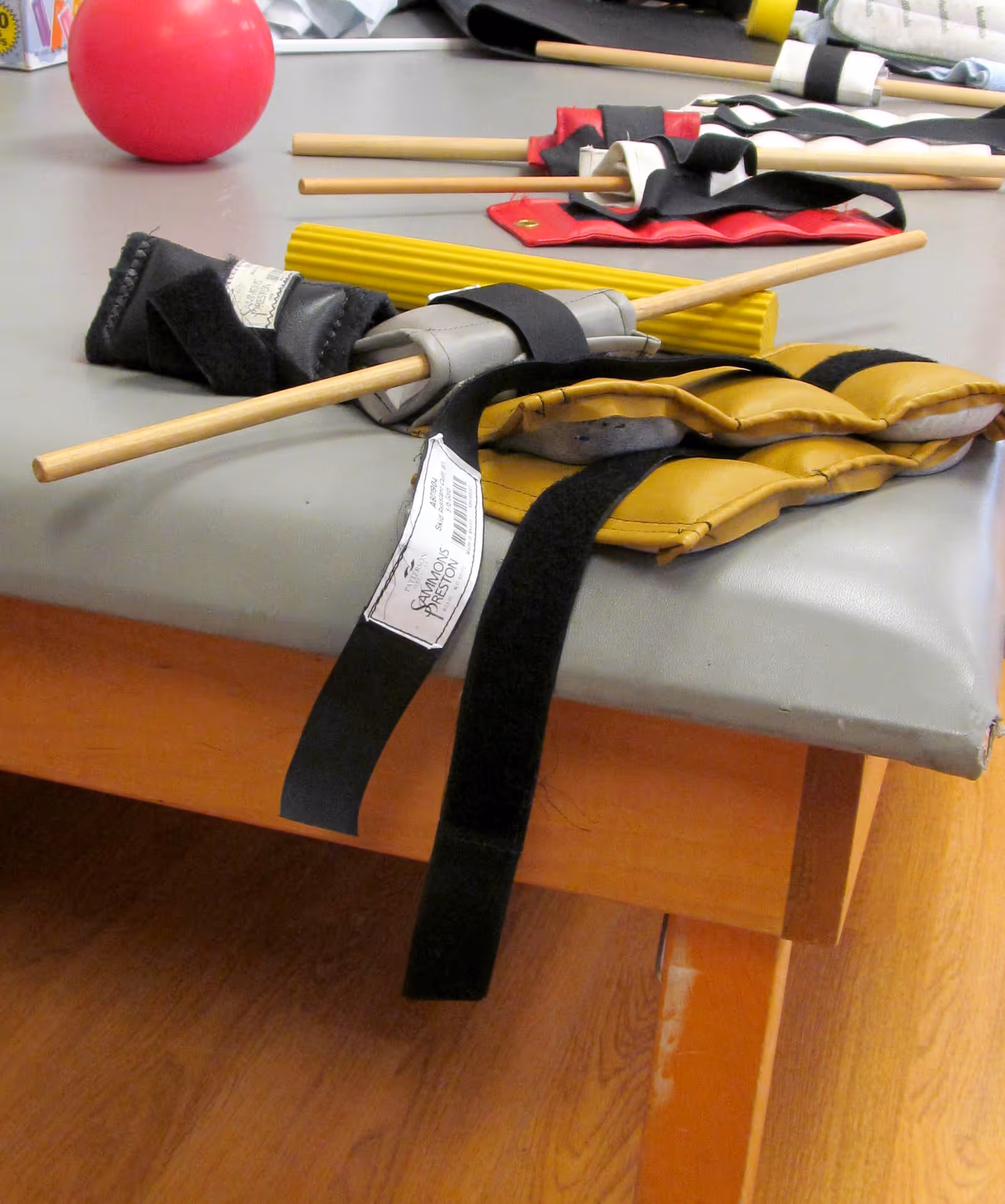 Close-up of various physical therapy equipment including padded straps with wooden rods, a yellow textured roller, and a red ball on a padded therapy table with a wooden frame and a hardwood floor beneath.