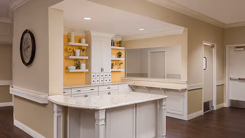 Interior view of a reception or common area with a white marble countertop desk, beige walls, wooden flooring, built-in shelves with decorative items and plants, and a large wall clock.
