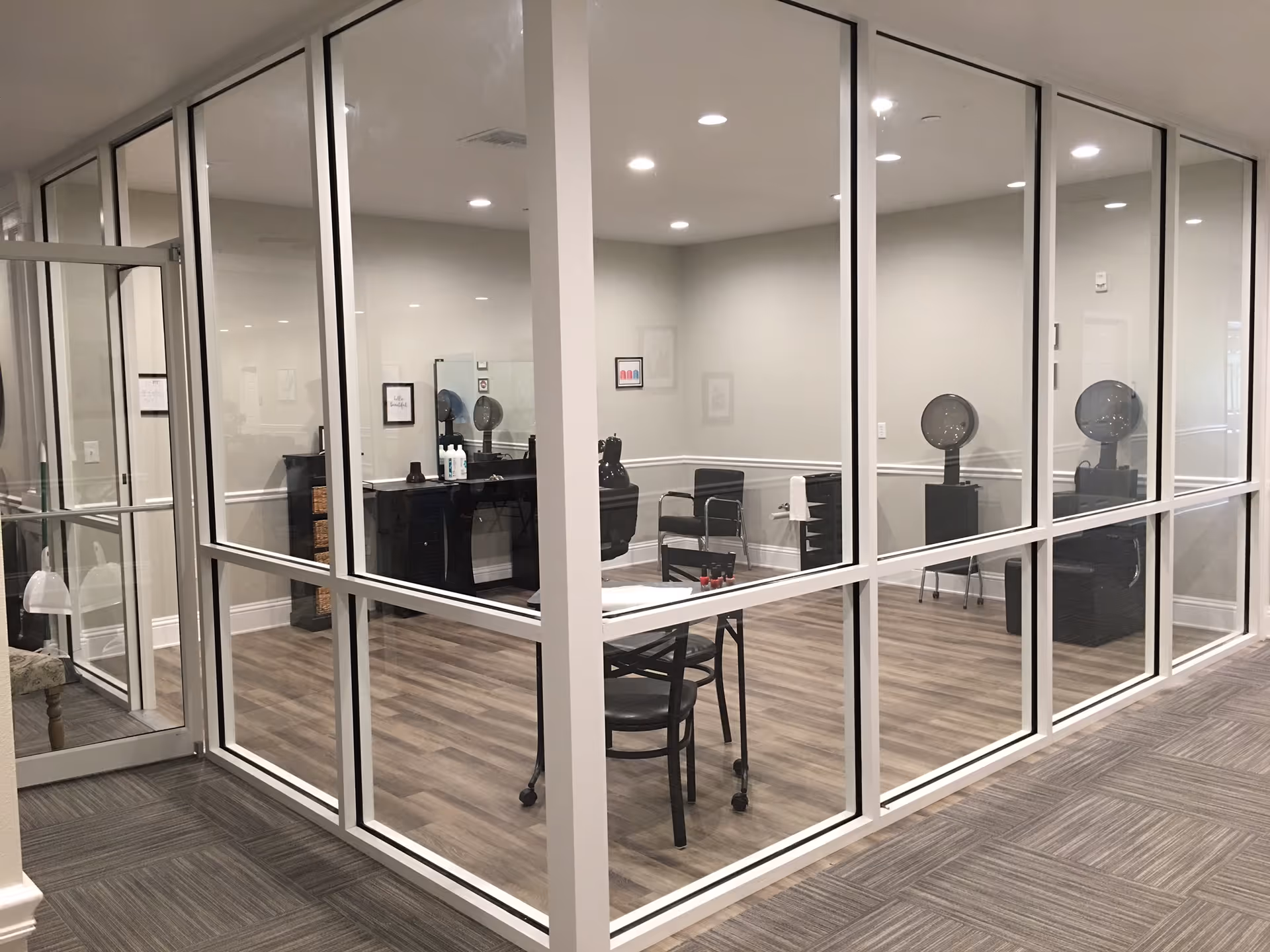 Glass-walled salon room in a senior living facility with styling chairs, hooded hair dryers, and a table.