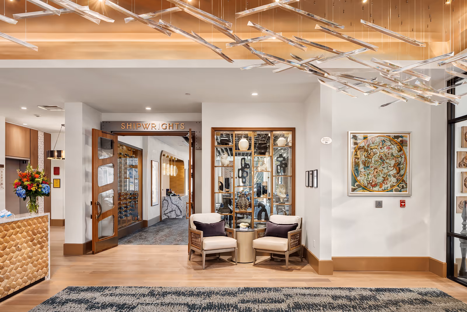 A modern and elegant interior space featuring a seating area with two cushioned chairs and a small round table between them. Behind the chairs is a decorative glass display with artistic objects. To the left, there is an open doorway with wooden doors labeled 'SHIPWRIGHTS' leading to another room with a reception desk and additional seating. The ceiling has a unique hanging light fixture with multiple rectangular elements. The walls are adorned with framed artwork, and a large floral arrangement is visible on the left side near a textured counter.