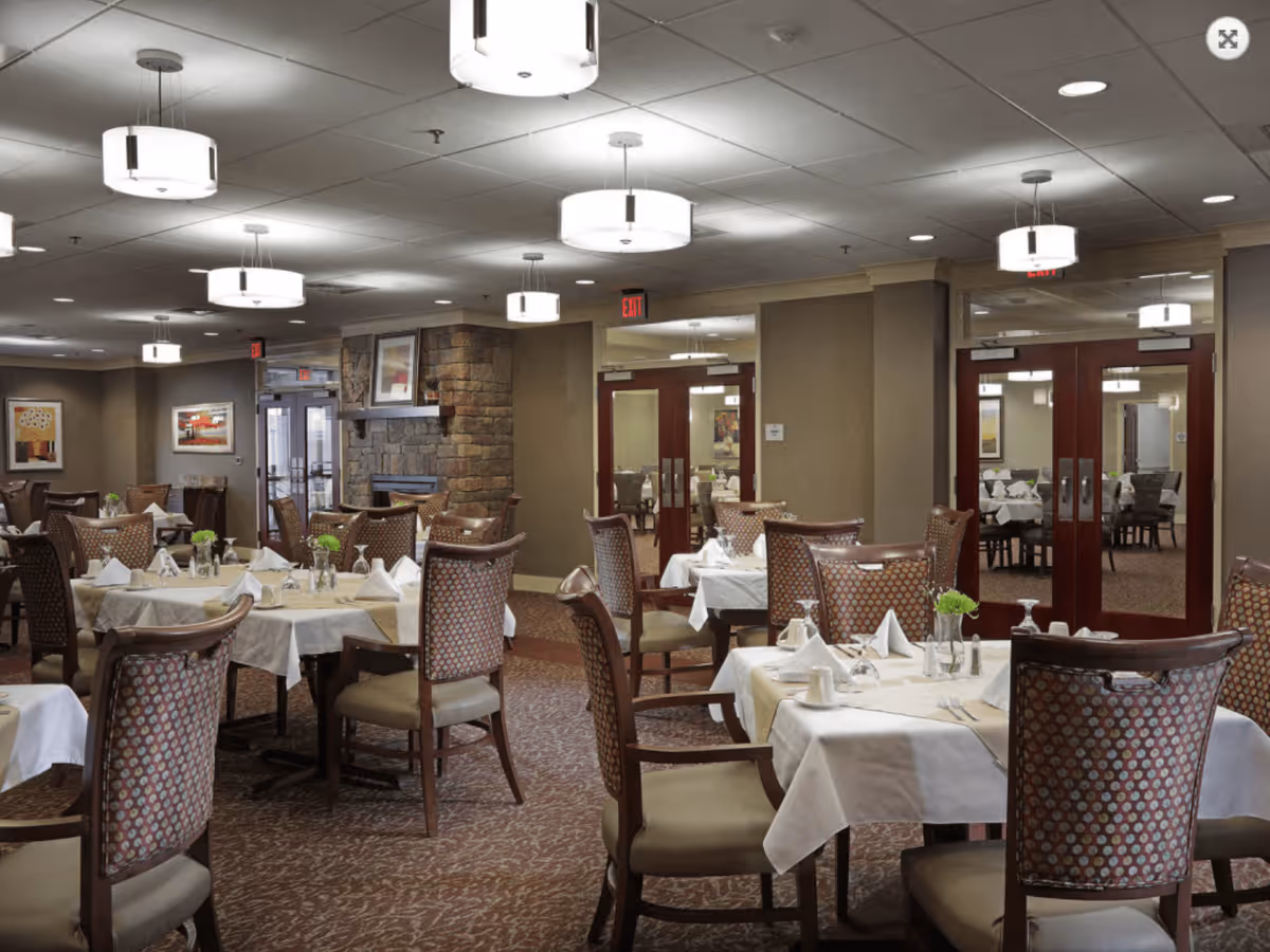 A spacious dining room in an assisted living center with multiple tables covered in white tablecloths, each set with folded napkins, glassware, and small green floral centerpieces. The room features patterned upholstered chairs, carpeted floors, warm lighting from ceiling fixtures, and a stone fireplace on one wall. Double wooden doors with glass panels lead to another dining area.