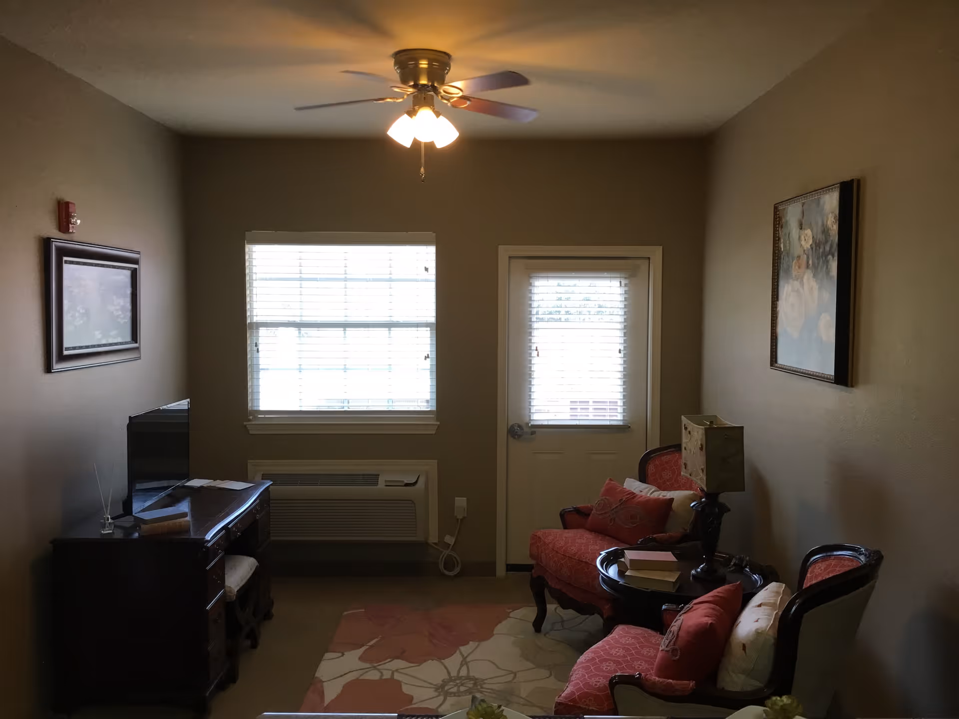 A cozy living room in a senior living facility with a ceiling fan and light fixture. The room has a window and a door with blinds, a wooden desk with a TV on it, two upholstered chairs with red cushions, a small round table with a lamp and books, and framed artwork on the walls. A floral area rug covers part of the floor.