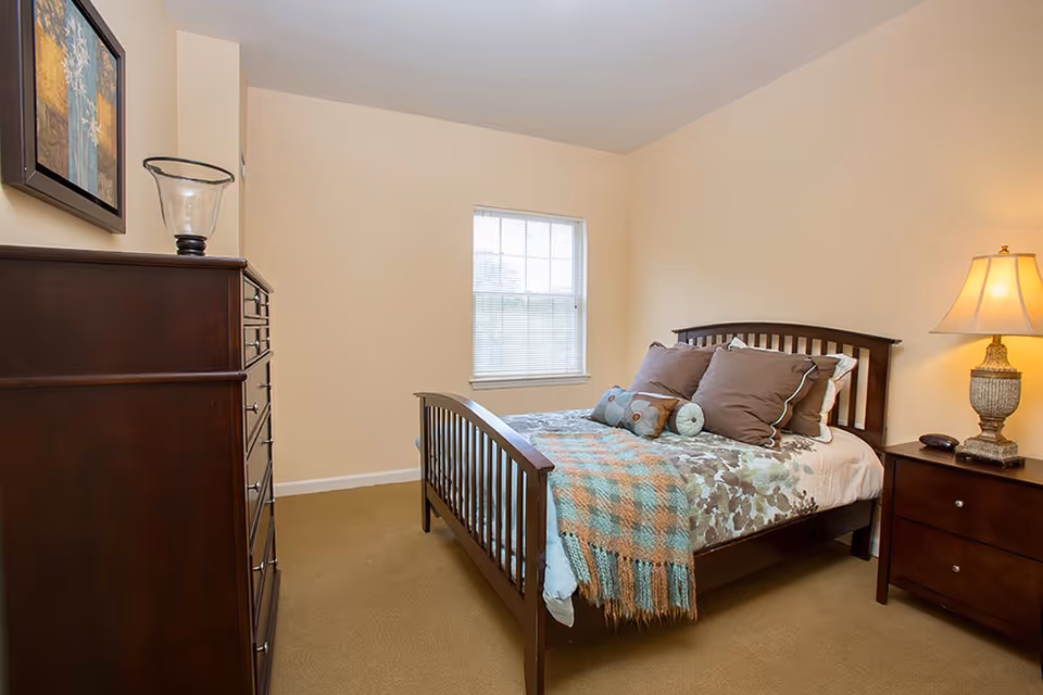 A cozy bedroom with a wooden bed frame, a bed made with floral and plaid bedding, several pillows, a wooden nightstand with a lamp, and a wooden dresser with a decorative glass vase. A window with blinds is centered on the far wall.