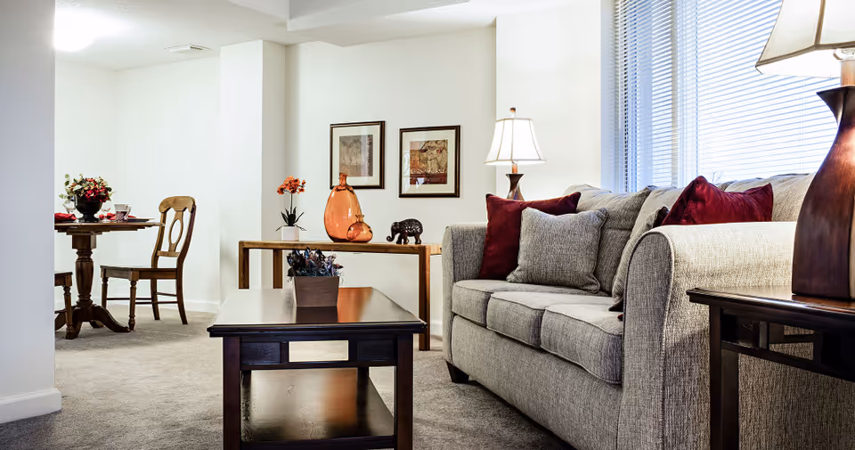 A cozy living room area with a beige sofa adorned with burgundy and gray pillows, a dark wooden coffee table with a small plant, a side table with a lamp, and a console table with decorative items including an orange vase and an elephant figurine. In the background, there is a small round dining table with two wooden chairs and a floral centerpiece. The room has light-colored walls and carpeted flooring, with large windows covered by blinds allowing natural light to enter.