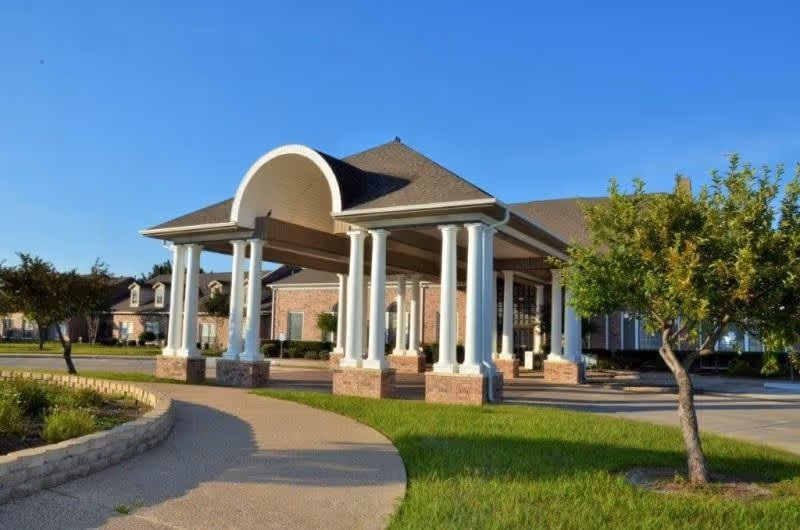 Entrance of a senior living facility with a covered driveway supported by white columns, surrounded by green grass, trees, and a clear blue sky.