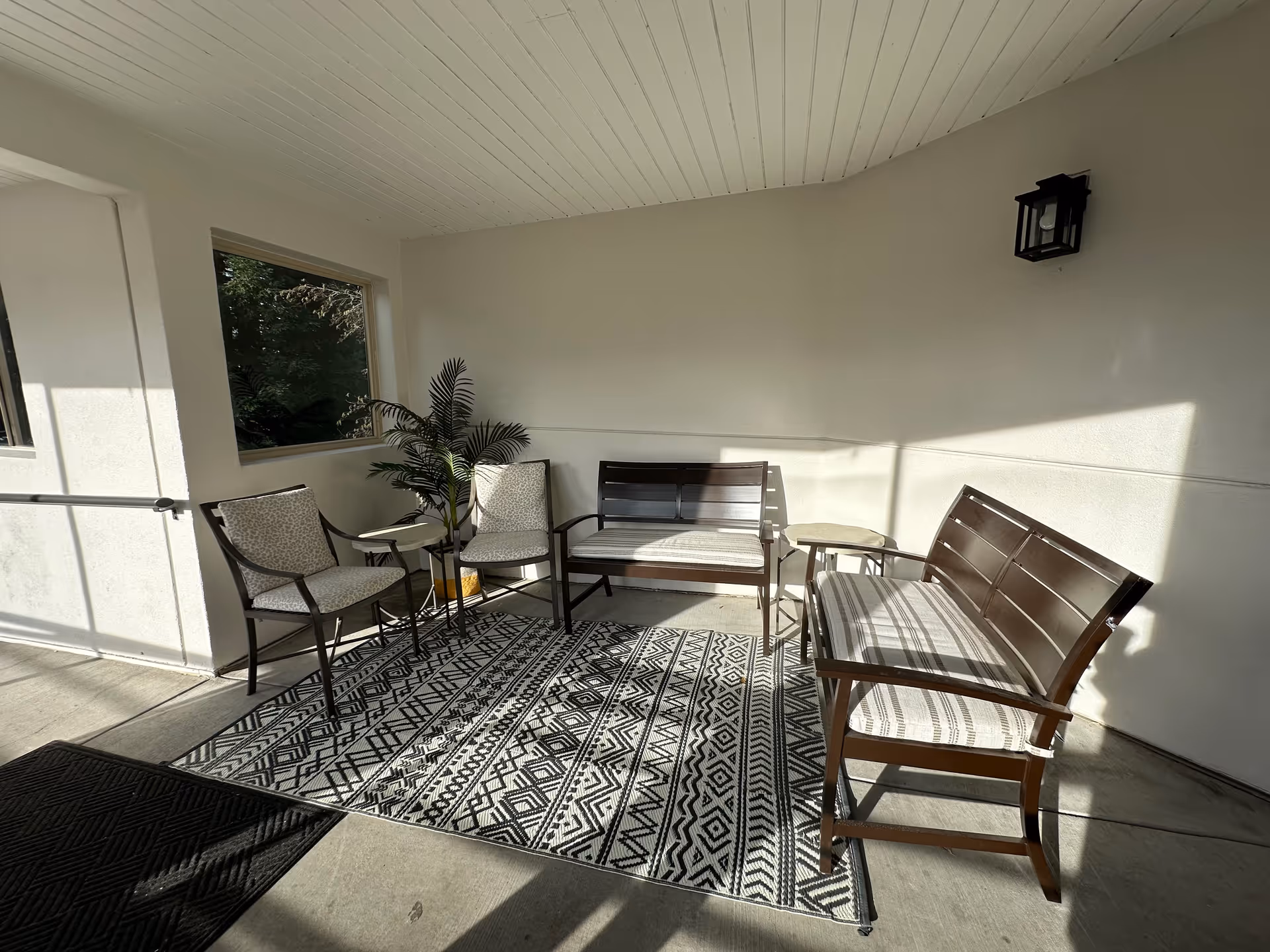Covered entry seating area with chairs, benches, side tables, a potted plant and a patterned rug lit by sunlight.