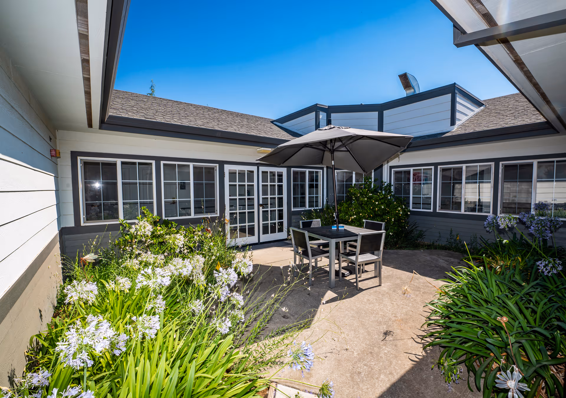 Outdoor courtyard area at Country Crest Post-Acute with a patio table and four chairs under a large umbrella, surrounded by flowering plants and bushes, with windows and doors of the building visible in the background under a clear blue sky.