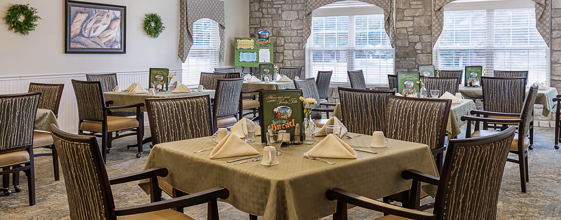 A dining room with multiple tables covered in beige tablecloths, each set with folded napkins, cups, and silverware. The room features large windows with white blinds and valances, stone accent walls, framed artwork, and small green wreaths on the walls. Chairs with patterned upholstery surround the tables.