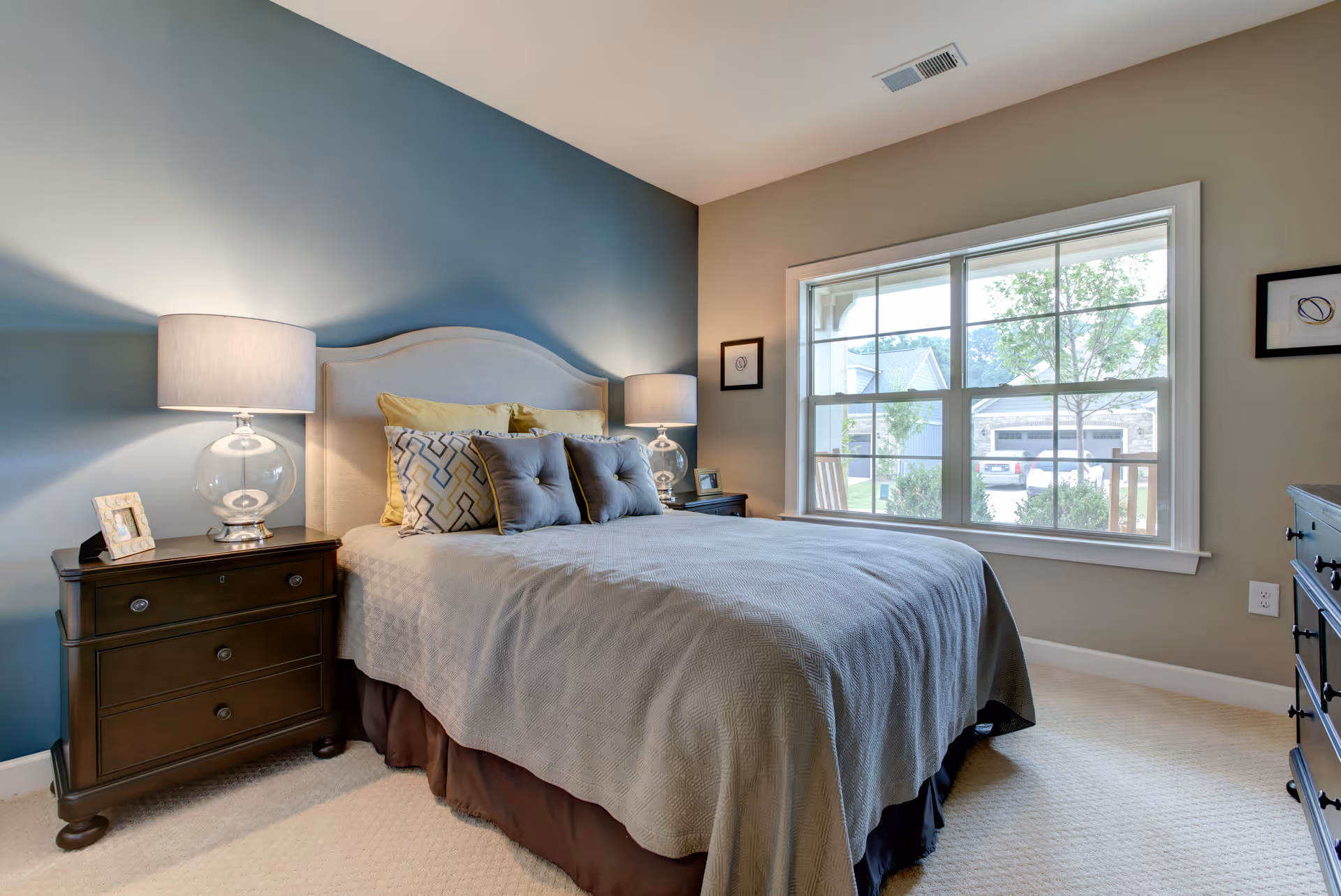 A cozy bedroom with a large bed featuring a beige upholstered headboard, multiple pillows, and a gray bedspread. There are two dark wooden nightstands on either side of the bed, each with a glass-based lamp. A large window on the right side lets in natural light and shows a view of houses and greenery outside. The walls are painted in two tones, with a darker blue accent wall behind the bed and a lighter beige on the other walls.