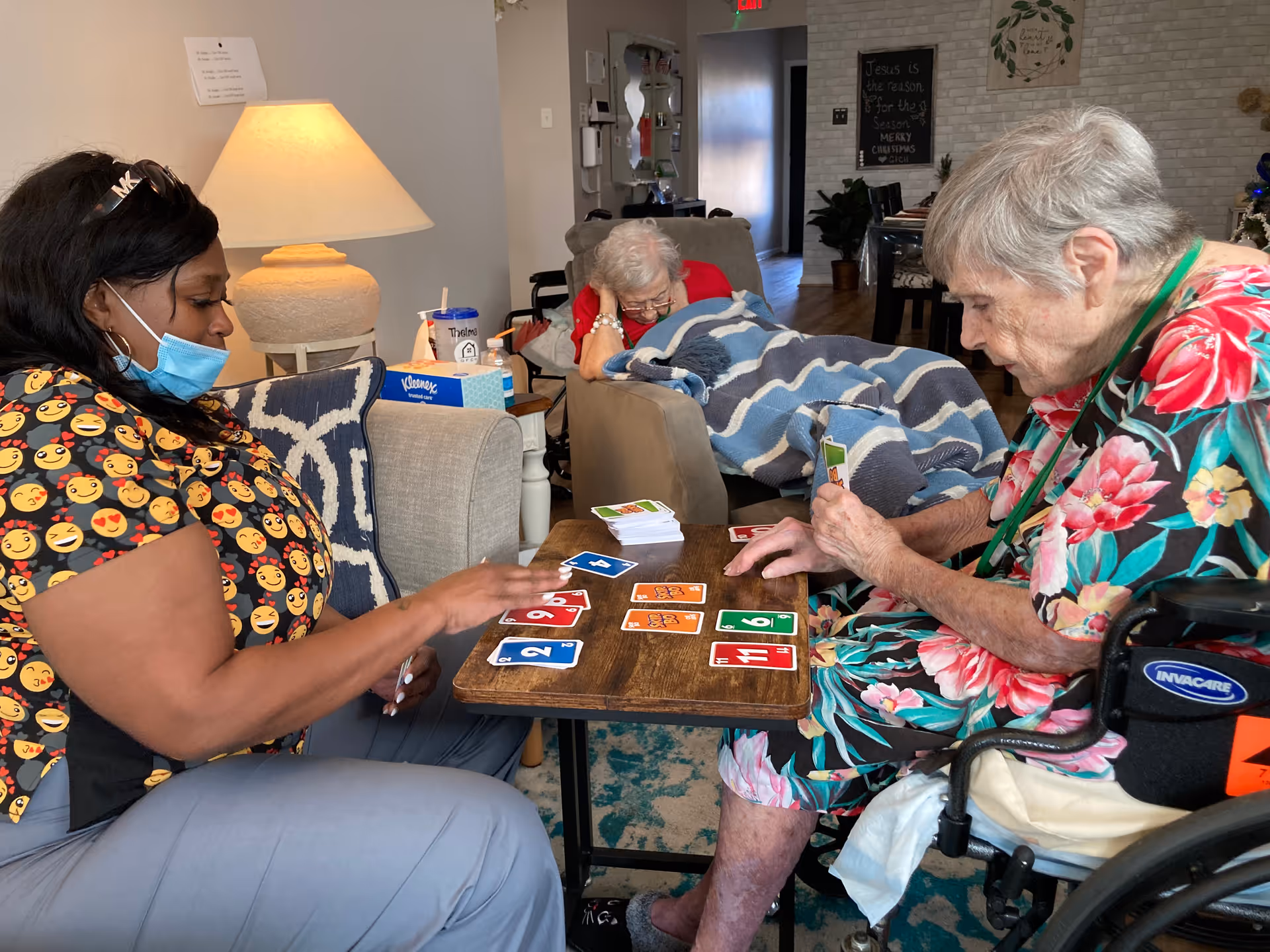 A caregiver wearing a mask and an emoji-patterned shirt plays a card game with an elderly woman in a floral dress seated in a wheelchair. Another elderly woman is resting on a couch covered with a blanket in the background. The setting is a cozy living room with a lamp, tissue box, and a sign on the wall.