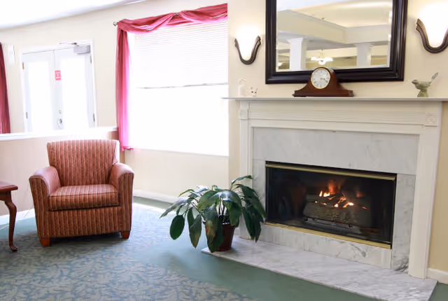 A cozy living room area with a lit fireplace surrounded by a white marble mantel. Above the fireplace is a large mirror and two wall sconces. To the left of the fireplace is a potted plant on the floor and a striped upholstered armchair near a window with pink curtains. The floor is carpeted with a patterned design.