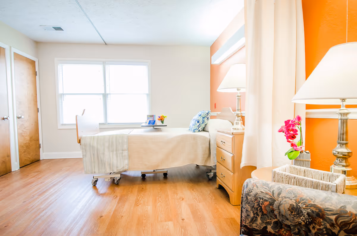 A bright and clean bedroom in a senior living facility with a single bed covered in beige bedding, a wooden nightstand with a lamp, a floral pillow, a window letting in natural light, a wooden door, and a floral-patterned armchair next to a small table with a lamp and a pink orchid.