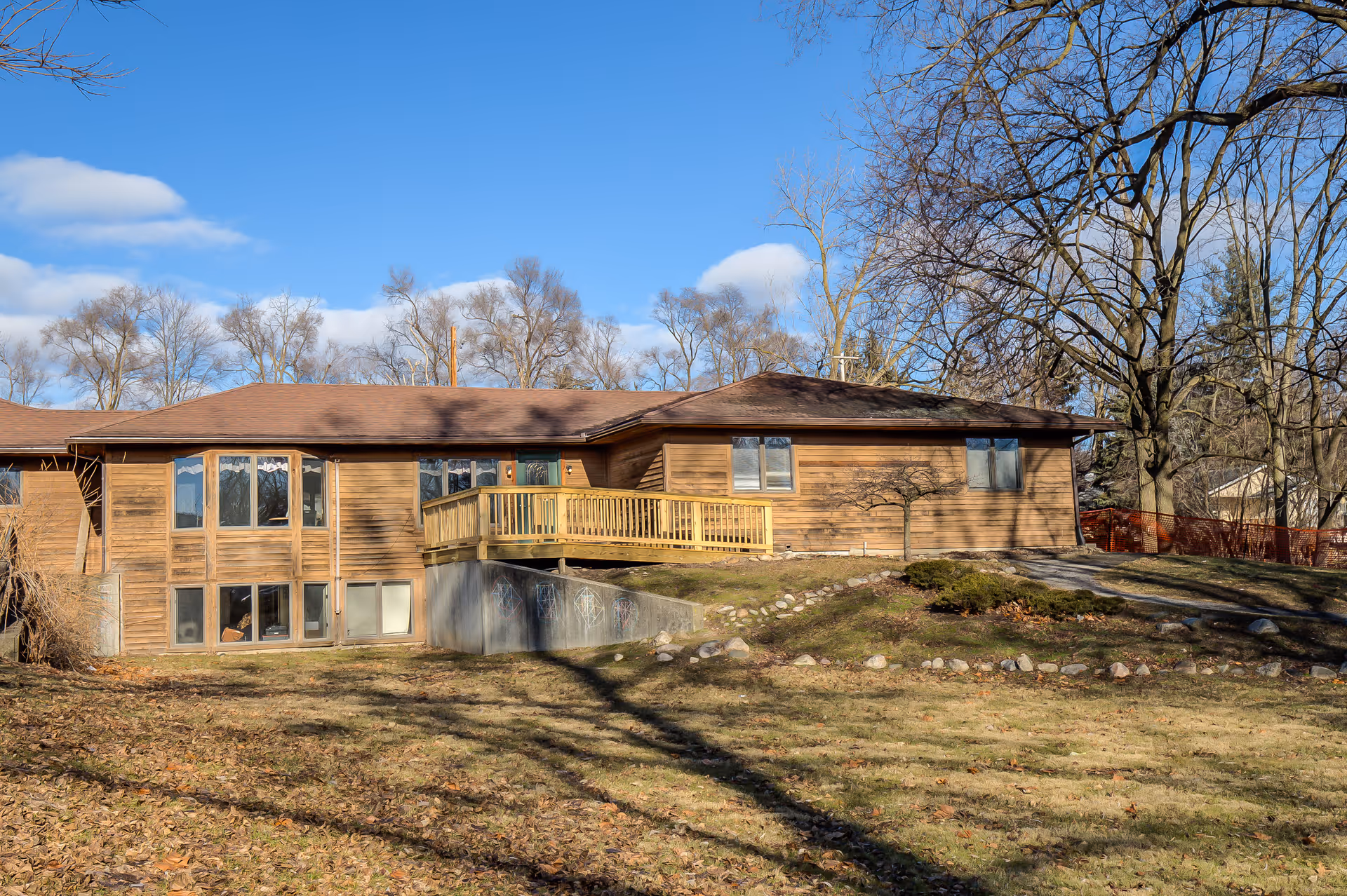 Exterior view of a single-story wooden building with a brown roof, large windows, and a small wooden deck. The building is surrounded by leafless trees and a grassy yard under a clear blue sky.