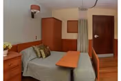 Small, tidy patient bedroom with a single bed, overbed table, wooden dresser and wardrobe, a wall lamp, and a privacy curtain.