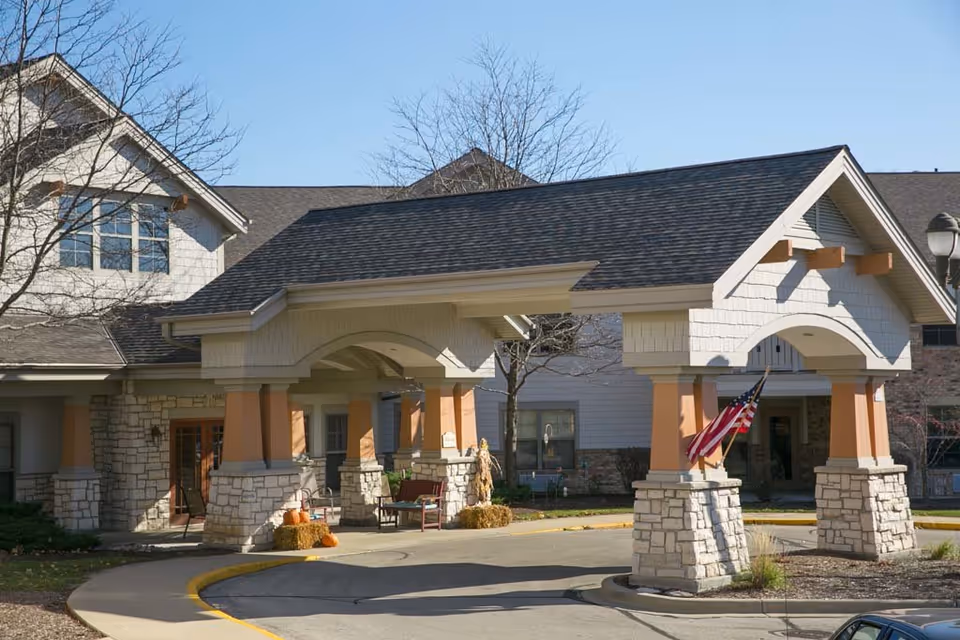 Entrance of a senior living facility with a covered driveway supported by stone and beige pillars, an American flag mounted on one pillar, some outdoor seating, and autumn decorations including pumpkins and hay bales. The building has a stone and siding exterior with a shingled roof and leafless trees nearby under a clear blue sky.