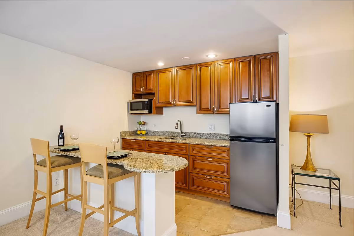 A small kitchen area with wooden cabinets, a granite countertop, a stainless steel refrigerator, a microwave, and a sink. There is a breakfast bar with two wooden chairs, each set with a plate and wine glass. A bottle of wine is placed on the counter. To the right, there is a small glass side table with a lamp on it.