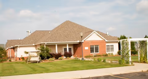 Single-story brick and siding building with a pitched roof, surrounded by a well-maintained lawn and landscaping, featuring a white pergola and a wooden swing in front of the building under a partly cloudy sky.